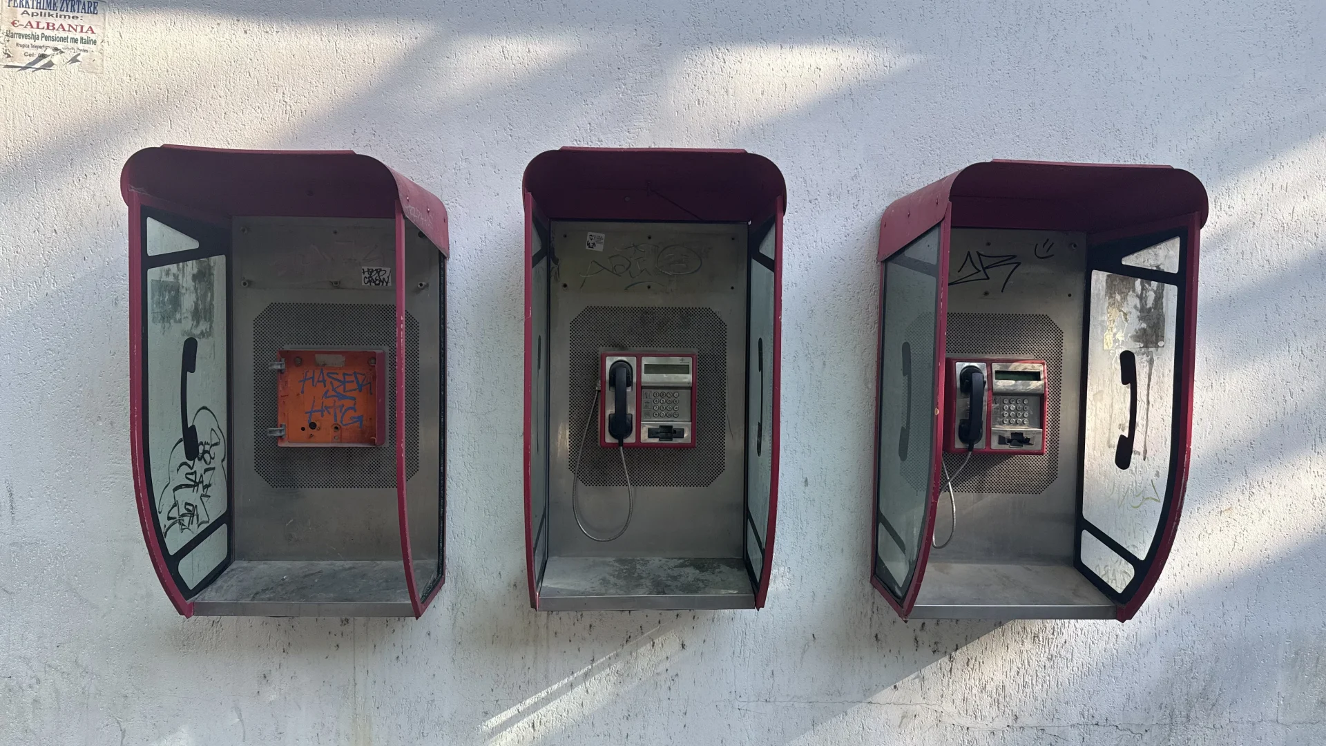 Een rij kleurrijke, verweerde oude telefooncellen langs een straat in Tirana, Albanië, met op de achtergrond gebouwen en voorbijgangers. Een knipoog naar de overstap van klassieke telefonie naar de moderne eSIM.