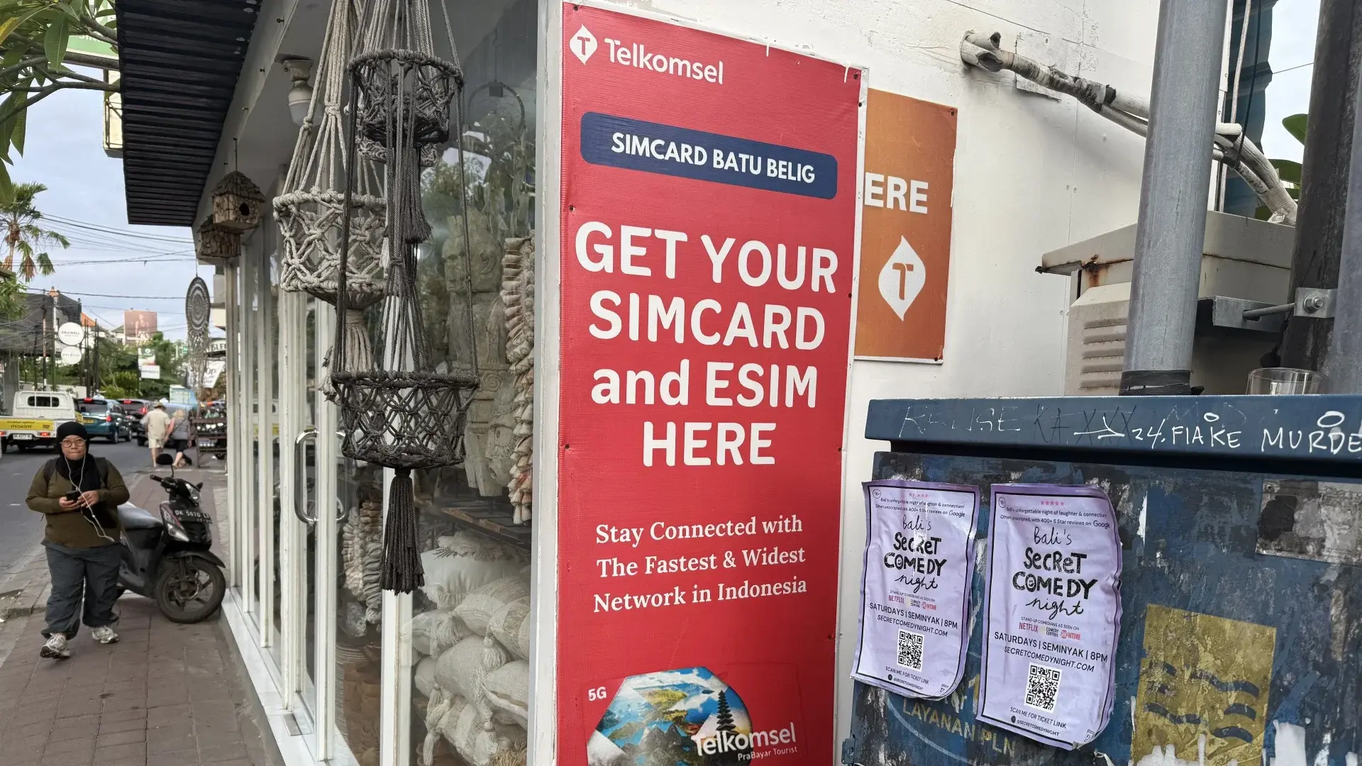Advertentie voor Telkomsel-simkaarten en eSIM's op een rode banner bij een winkeltje in Batu Belig, nabij Seminyak op Bali, met een voorbijlopende vrouw en een geparkeerde scooter.