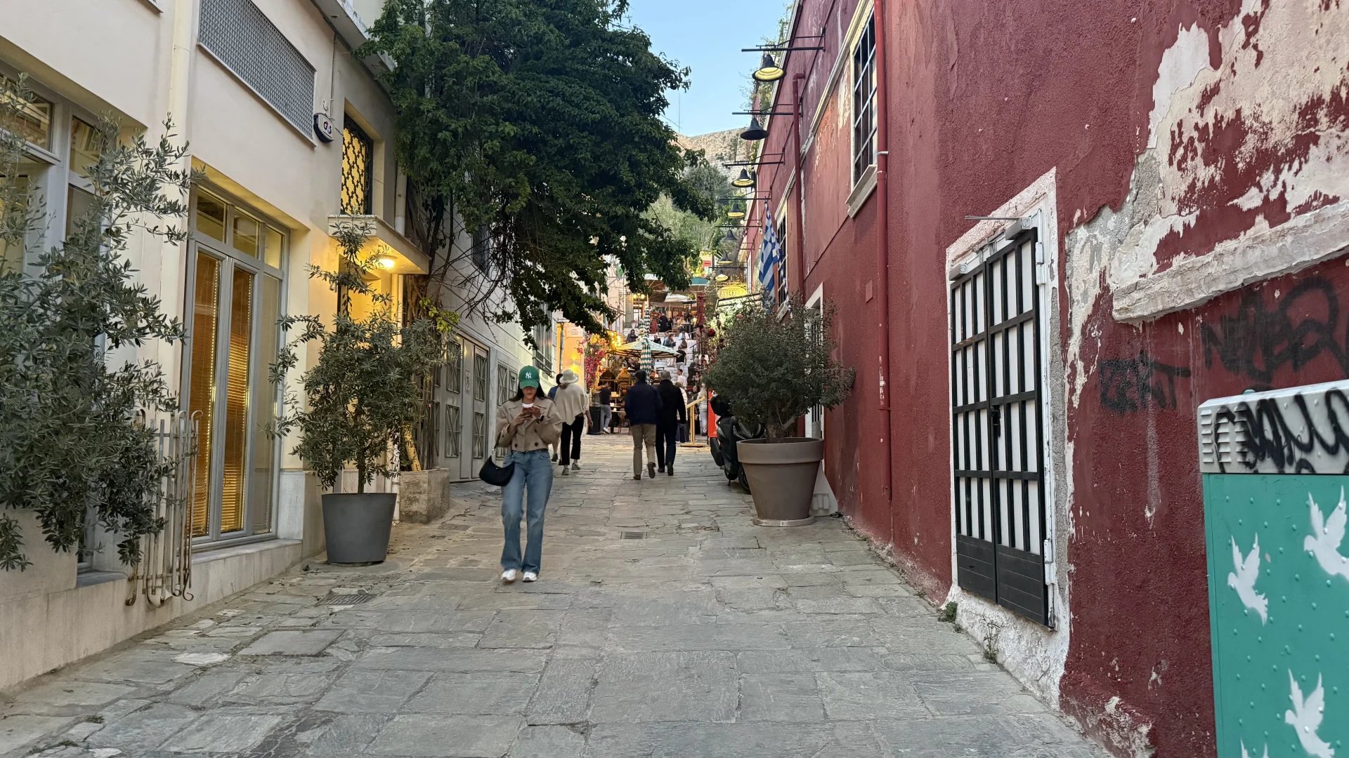 Een sfeervolle, met kasseien bestrate hellende straat in de wijk Plaka in Athene. Links een lichtgekleurd gebouw met grote ramen en olijfbomen in potten; rechts een opvallende verweerde donkerrode muur met graffiti. Voetgangers lopen bergop richting terrasjes en weelderig groene bomen onder een heldere hemel.