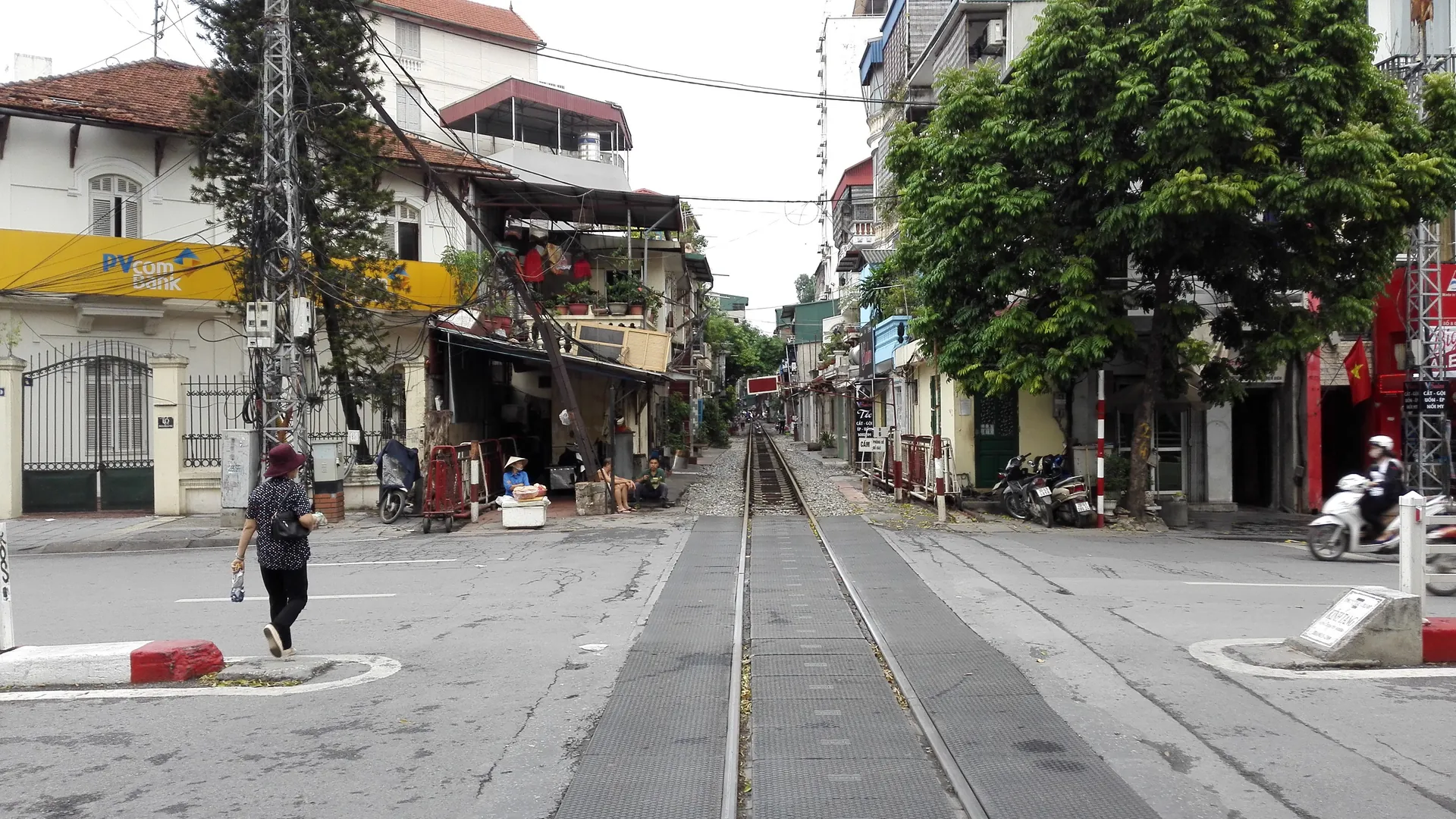 Een spoorlijn die dwars door een straatkruising in Hanoi loopt, met winkelhuizen, een bank en een scooter aan weerszijden van de rails