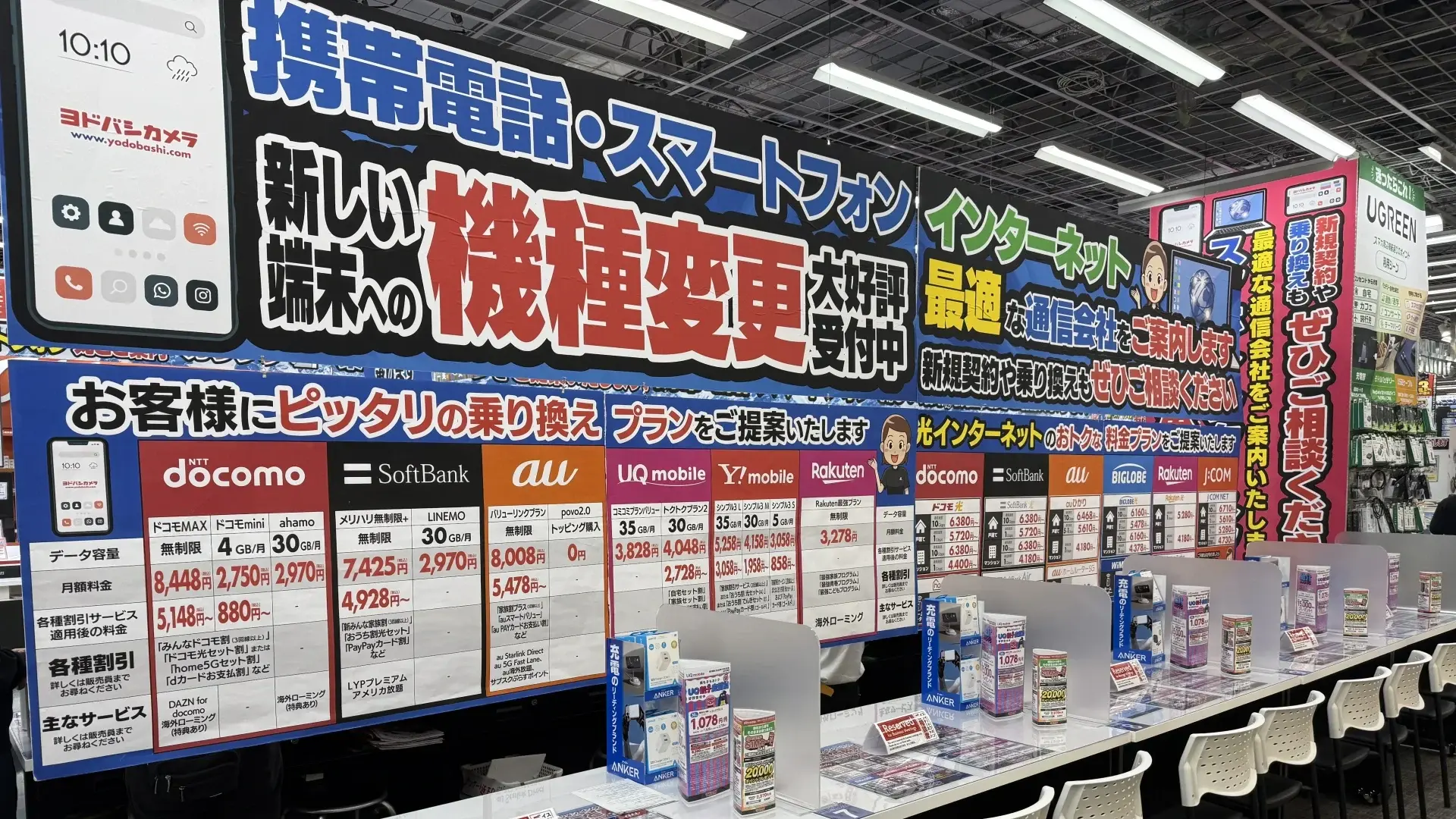 Een mobiele servicebalie in een Yodobashi Camera-winkel met grote kleurige banners en prijslijsten voor NTT Docomo, SoftBank, au en Rakuten Mobile
