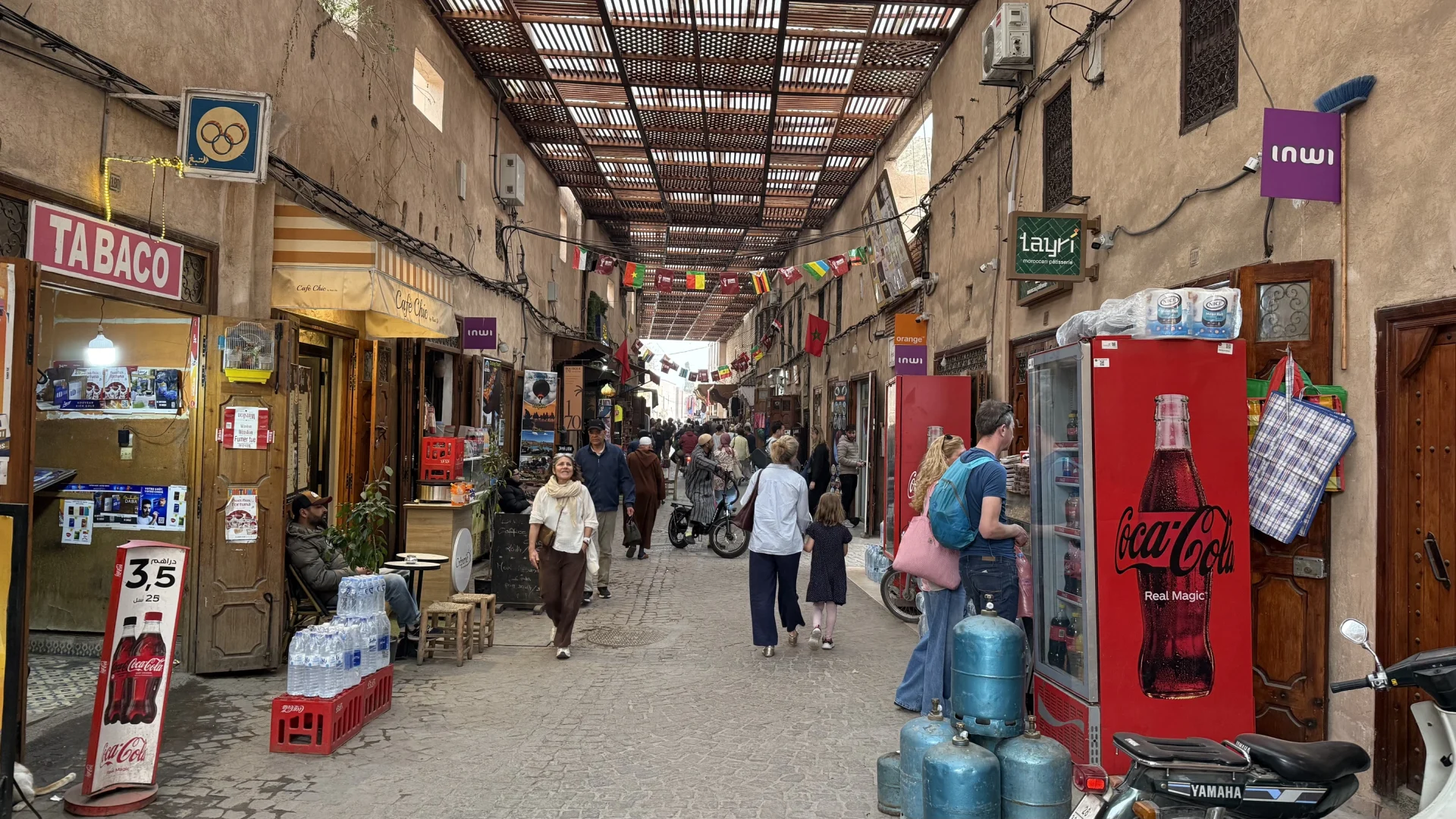 Overdekt marktsteegje in Marrakesh met kleine winkeltjes, telecom-uithangborden (Inwi en Orange), een Coca-Cola-koelkast en voorbijgangers.