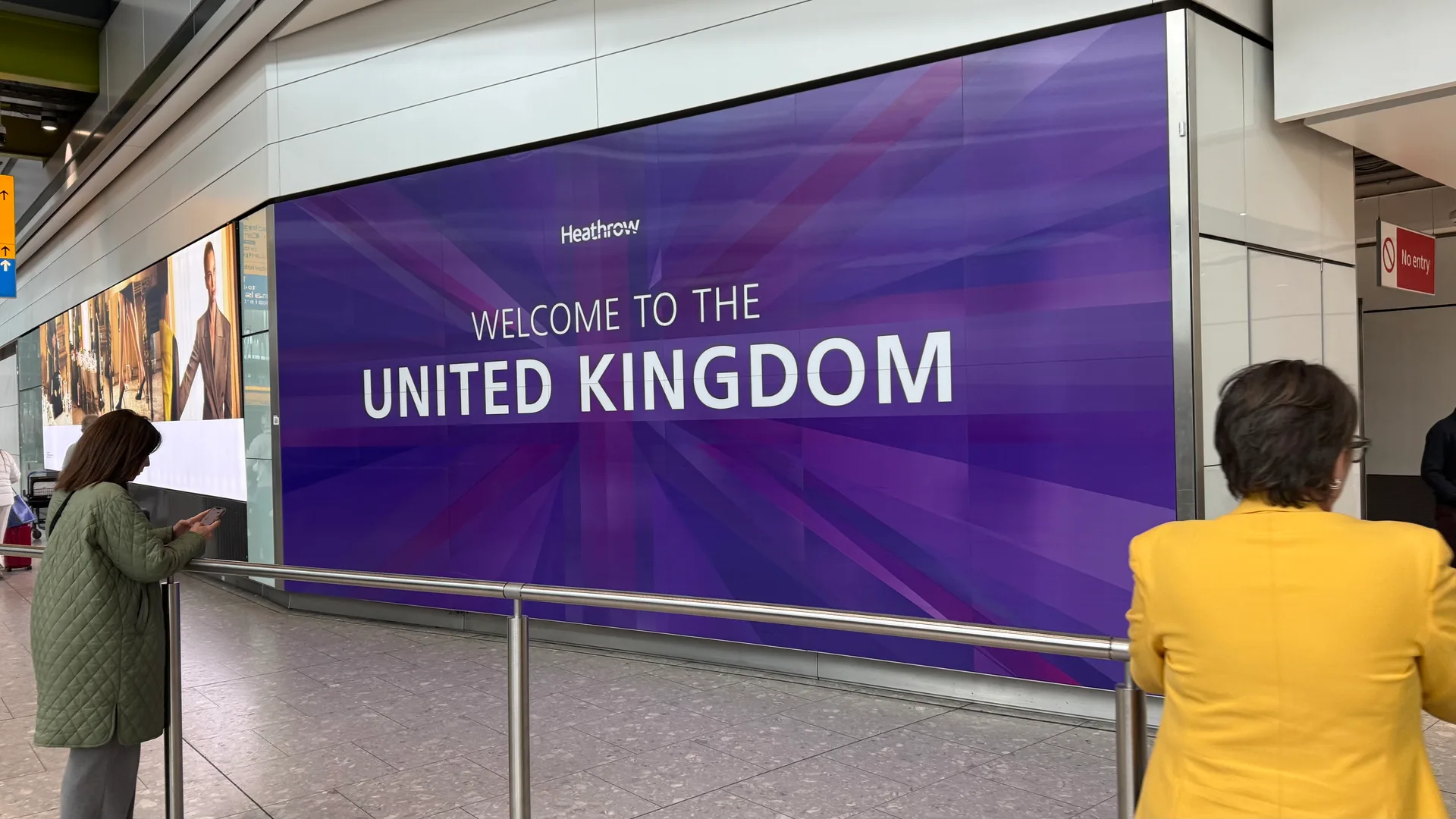 Groot paars "Welcome to the United Kingdom"-scherm in de aankomsthal van London Heathrow, met een reiziger die haar telefoon checkt