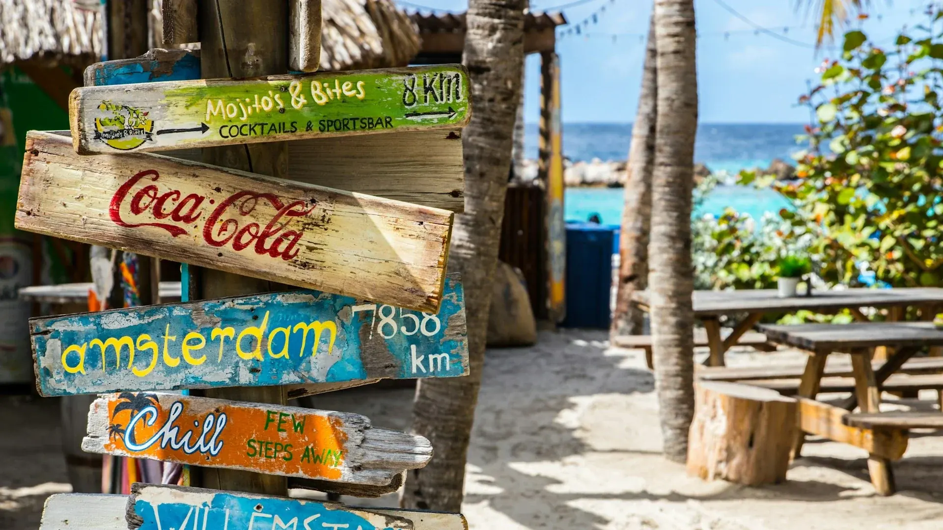 Een kleurrijke, rustieke houten wegwijzer op een zonnig strand op Curaçao, met beschilderde bordjes die wijzen naar bestemmingen zoals Amsterdam, Willemstad en lokale beachclubs.
